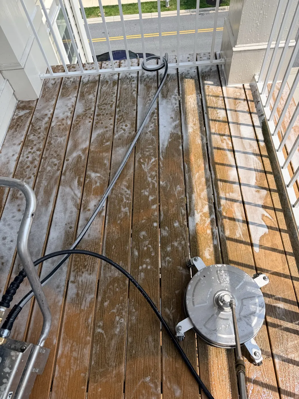A surface cleaner and pressure hose on a wood deck mid-wash, cleaning solution lifting the grime off the boards.