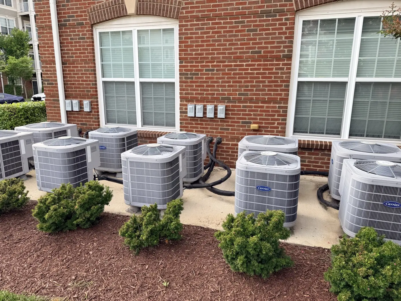 A row of HVAC condenser units beside clean brick wall and windows on a residential building.